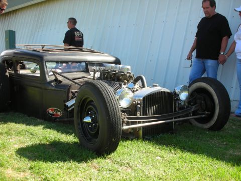 1998 Ford Crown Victoria - very low flat-black chopped rat rod sedan with an exposed chromed V8 and Black Flag plaque sits on grass as men look on.