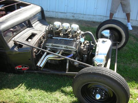 1998 Ford Crown Victoria - Detail of a flat-black rat rod's exposed V8 with three carburetors, headers and Black Flag plaque on a tube chassis.
