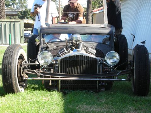 1998 Ford Crown Victoria - Head-on view of a low flat-black rat rod showing its vertical grille, twin round headlights and exposed engine as people gather behind.