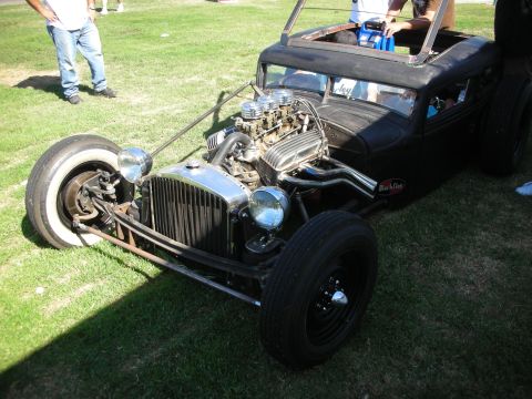 1998 Ford Crown Victoria - low flat-black channeled rat rod with an exposed tri-power V8 and a wide whitewall front tire sits on grass at a show.