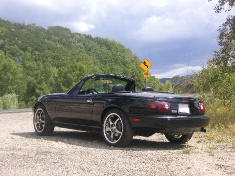 1996 Mazda Miata - A black convertible sports car is parked on a gravel road, with a winding road sign in the background.