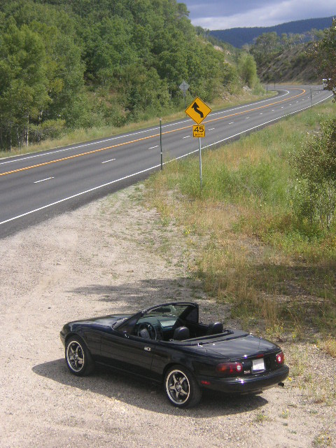 1996 Mazda Miata - A black 1996 Mazda Miata convertible sits on a dirt pull-off beside a scenic, winding highway.