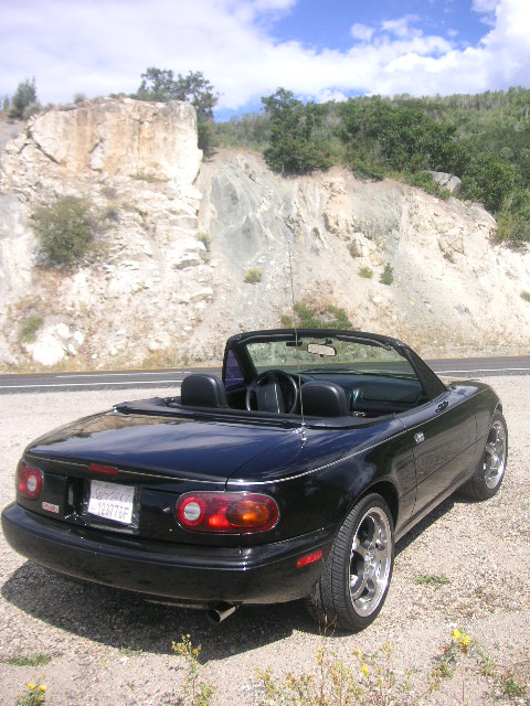 1996 Mazda Miata - Black 1996 Mazda Miata convertible parked on a roadside with a rocky hill behind it.