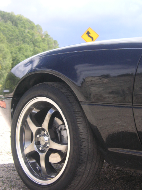 1996 Mazda Miata - A dark blue car's side profile is shown, with a focus on the front wheel and fender, under a bright sky.