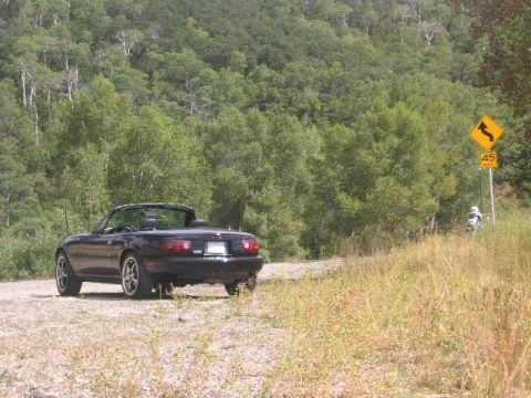 1996 Mazda Miata - A black 1996 Mazda Miata convertible is parked on a dirt road, with a wooded hillside and a speed limit sign in the background.