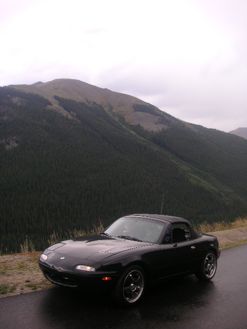 1996 Mazda Miata - Black 1996 Mazda Miata parked on a wet mountain road with pine-covered slopes in the background.