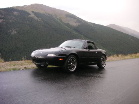 1996 Mazda Miata - A black convertible sports car is parked on a wet road with a mountainous, forested landscape behind it.