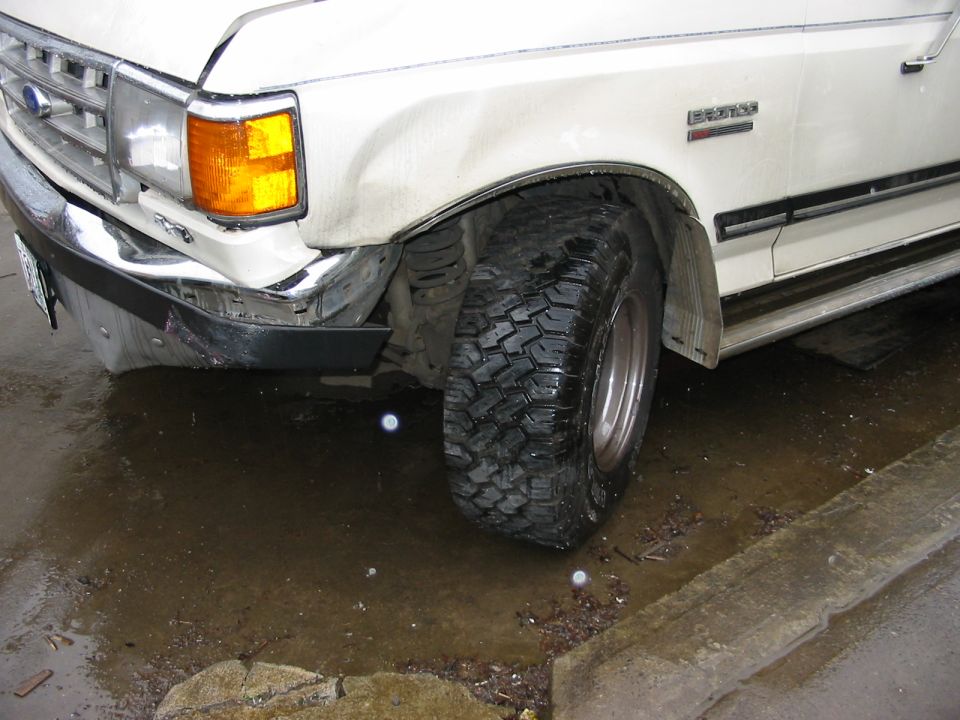 Front corner of a white 1980s Ford F-Series truck with a damaged bumper and mud-terrain tire