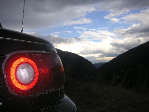 1996 Mazda Miata - The rear of a dark-colored car is visible with its tail light illuminated, set against a dramatic cloudy sky and mountainous landscape.