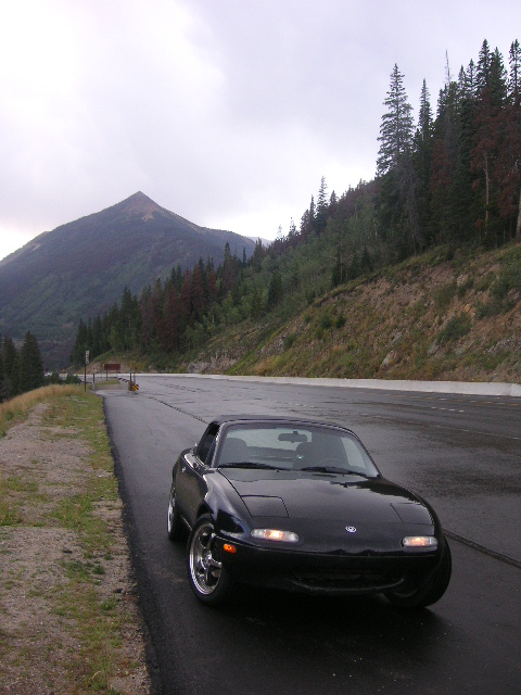 1996 Mazda Miata - A black 1996 Mazda Miata is seen on a scenic mountain road under an overcast sky.