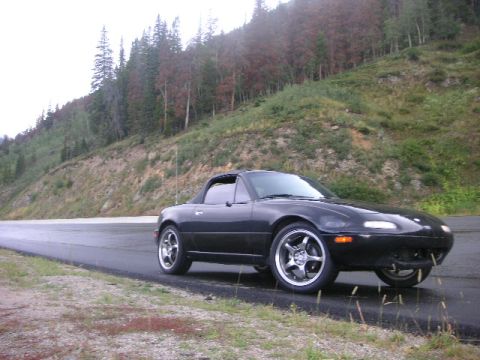 1996 Mazda Miata - A black convertible sports car is parked on the side of a wet road, with a wooded hillside in the background.