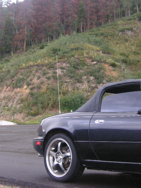 1996 Mazda Miata - Dark colored Mazda Miata parked on a paved surface with a wooded hillside in the background.