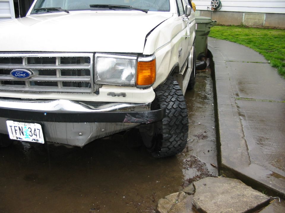 Front corner of a white 4x4 Ford truck with damaged bumper parked on a wet driveway
