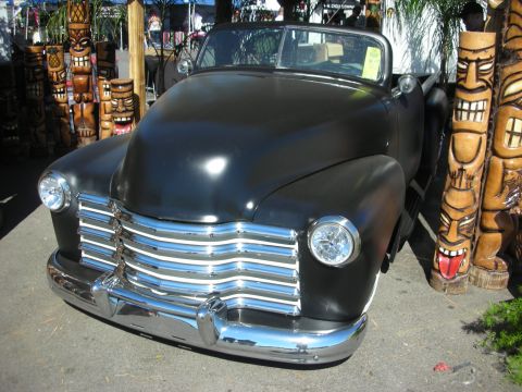 1998 Ford Crown Victoria - flat-black 1940s Cadillac with a heavy chrome grille sits between hand-carved tiki totem poles at an outdoor venue.