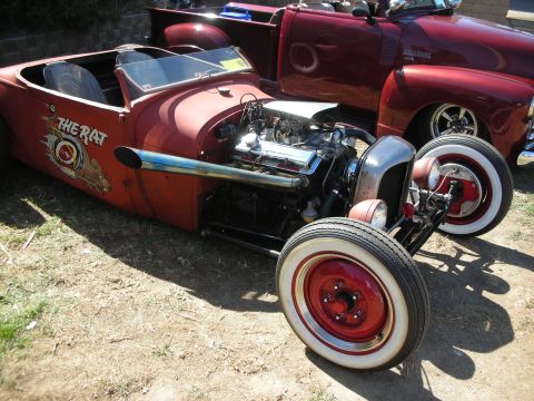 1998 Ford Crown Victoria - weathered rust-patina hot rod roadster lettered THE RAT with an exposed V8 and red steel wheels sits on grass.