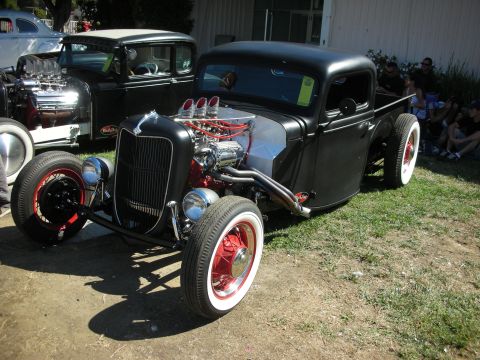 1998 Ford Crown Victoria - flat-black hot rod pickup with a chromed tri-power V8, red steel wheels and whitewall tires sits on grass at a show.