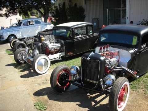 1998 Ford Crown Victoria - Several black chopped hot rods and coupes with exposed chrome engines line up on grass beside a building at a car show.