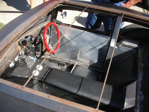1998 Ford Crown Victoria - Unfinished rat rod interior showing a red steering wheel, floor shifter and black bench seats inside a stripped cabin.