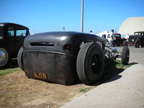 1998 Ford Crown Victoria - heavily lowered flat-black rat rod sits on grass at a car show, its rear deck wearing a yellow license plate.