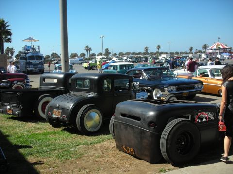 1998 Ford Crown Victoria - Several lowered flat-black rat rods line up along a paved lot at a seaside car show with palms and a tent in the background.