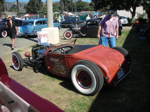 1998 Ford Crown Victoria - orange weathered roadster lettered THE RAT sits on grass at a car show as a man in plaid leans over to inspect it.