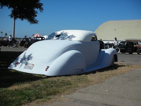 1998 Ford Crown Victoria - primer-white heavily chopped and lowered custom coupe sits on grass near a hangar at a car show with hot rods behind.