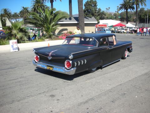 1998 Ford Crown Victoria - flat-black lowered late-1950s Ford sedan with round taillights cruises a paved lot lined with palms at a car show.