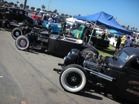 1998 Ford Crown Victoria - row of black rat rods with exposed V8 engines and whitewall tires lines a lot beside a blue canopy at a busy car show.