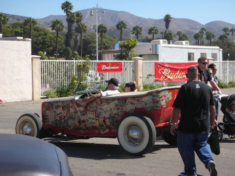 1998 Ford Crown Victoria - red open roadster hot rod with collage-printed bodywork and whitewall tires drives past spectators at a car show.