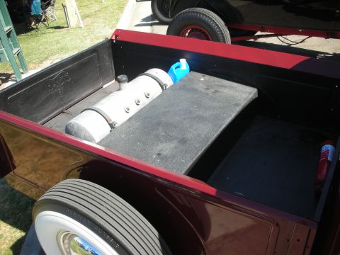 1998 Ford Crown Victoria - bed of a maroon hot rod pickup holds a polished cylindrical tank and a black storage box, with a whitewall tire below.