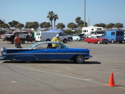 1998 Ford Crown Victoria - blue lowered vintage hardtop coupe sits in a parking-lot car show with vans, RVs and palm trees in the background.