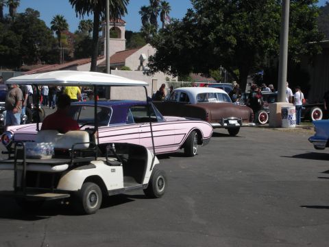 1998 Ford Crown Victoria - pink lowered custom car parked near a white golf cart and a brown coupe at a sunny outdoor car show.