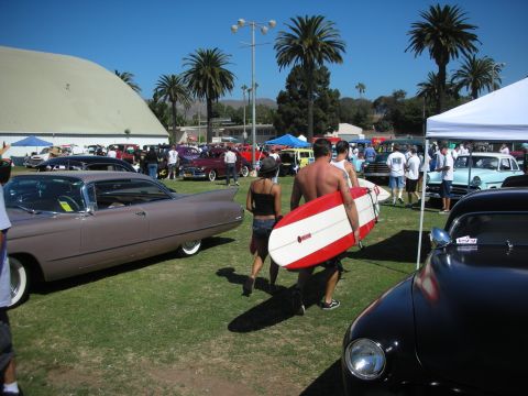 1998 Ford Crown Victoria - Two people carry a red-and-white surfboard between rows of custom cars and tents under palm trees at a car show.