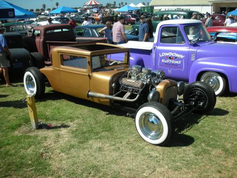 1998 Ford Crown Victoria - gold chopped hot rod with exposed engine and whitewalls parked beside a purple Lowdown Kustoms pickup at a show.