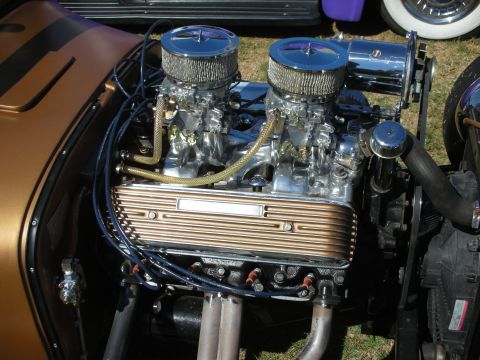 1998 Ford Crown Victoria - polished dual-carburetor V8 engine with finned valve covers and chrome air cleaners sits in a copper-colored hot rod at an outdoor car show.