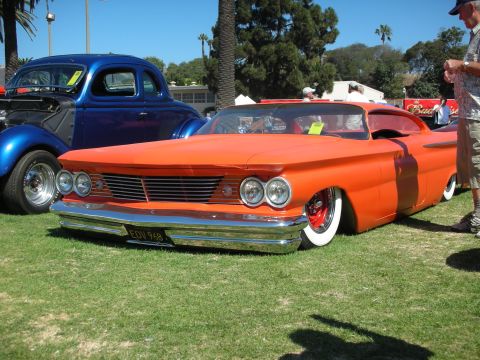 1998 Ford Crown Victoria - slammed satin-orange custom coupe with quad headlights and whitewall tires sits beside a blue coupe on a grassy car-show field.