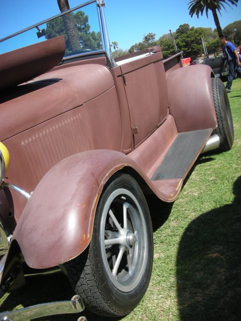 1998 Ford Crown Victoria - Low-angle view of the rear fender, tall tire and five-spoke chrome wheel of a primer-brown vintage roadster pickup under a blue sky.