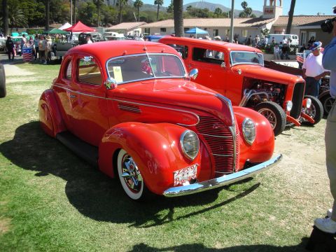 1998 Ford Crown Victoria - glossy red 1930s-style Ford coupe hot rod with chrome wheels and whitewalls sits on grass among other rods at an outdoor show.