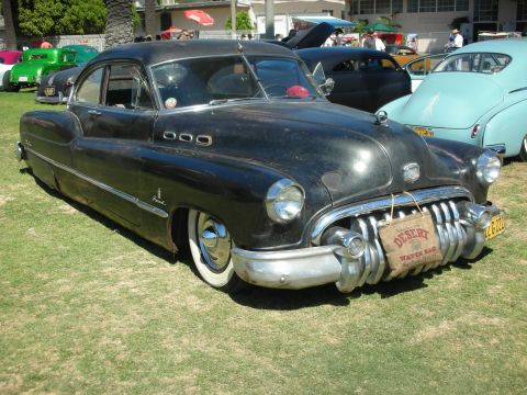1998 Ford Crown Victoria - weathered black 1950 Buick custom lead sled sits low on grass with a vintage Desert water bag hung from the chrome bumper.