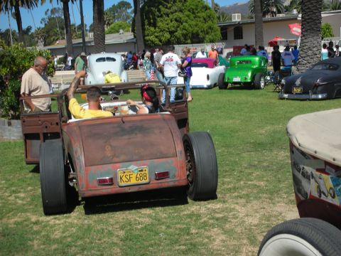 1998 Ford Crown Victoria - rusty patina rat-rod pickup carrying two occupants drives across a grassy show field past parked hot rods and a green coupe.
