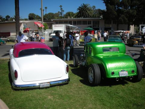 1998 Ford Crown Victoria - white custom car with red roof parked beside a metallic-green 1932-style five-window hot rod coupe at a busy outdoor car show.