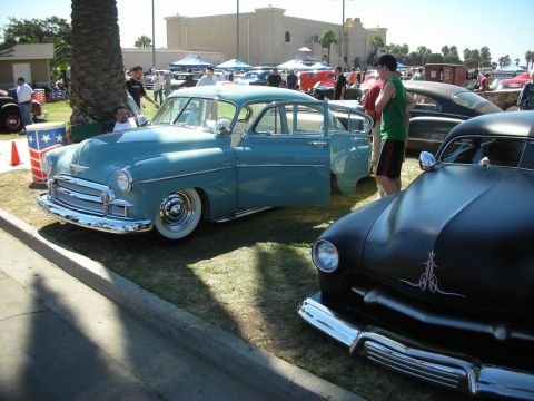 1998 Ford Crown Victoria - pale-blue lowered fifties sedan sits next to a flat-black custom with pinstriping in a crowded outdoor car show lot.