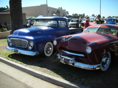 1998 Ford Crown Victoria - blue fifties pickup with a custom bullnose grille parks beside a maroon chopped lead-sled custom on a palm-lined show lot.