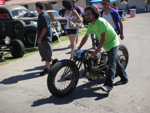 1998 Ford Crown Victoria - man in a green T-shirt rolls a vintage board-track-style motorcycle across the pavement past parked hot rods at a show.