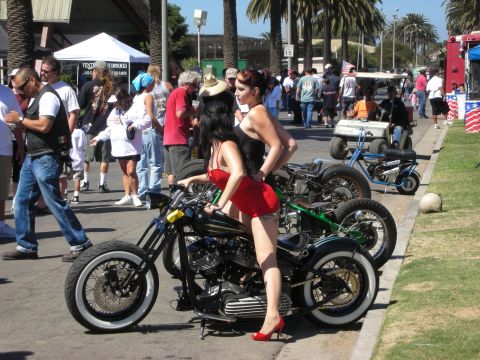 1998 Ford Crown Victoria - woman in a red dress and heels poses astride a black hardtail chopper among a row of bikes on a busy outdoor show midway.