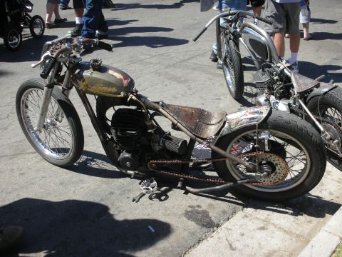1998 Ford Crown Victoria - weathered bare-metal hardtail bobber with an Indian-tank logo and rusty drag pipes sits low at a curb among other bikes.