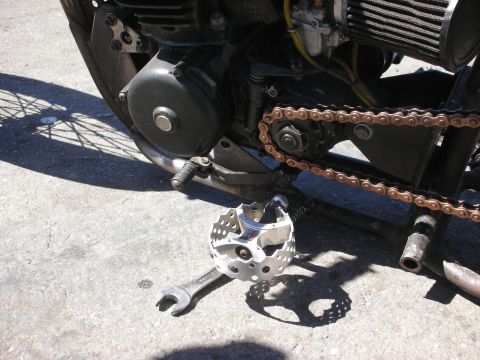 1998 Ford Crown Victoria - Detail of a bobber's engine, rusty drive chain and a skull-shaped polished foot peg, with a wrench lying on the pavement.