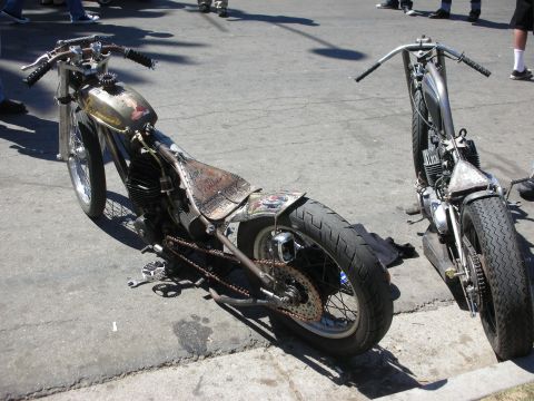 1998 Ford Crown Victoria - Two rough bare-metal hardtail bobbers with fat rear tires and patina tanks sit nose-to-nose against a curb at a bike show.