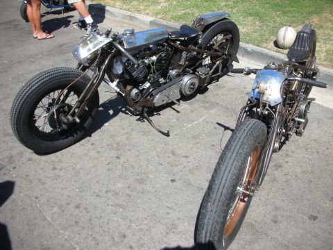 1998 Ford Crown Victoria - Two stripped bare-metal hardtail bobbers, one silver and one blue-tank, sit angled on the pavement at a motorcycle show.