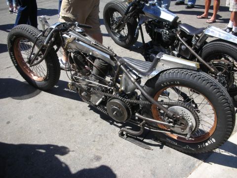 1998 Ford Crown Victoria - stripped bare-metal hardtail bobber shows its exposed V-twin engine, drive chain and a rear tire marked 4.50-18 Olympic.